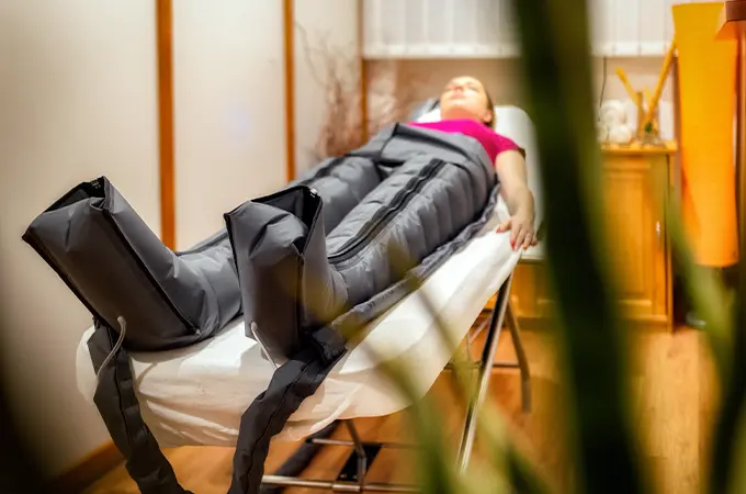 Kobieta relaksuje się na stole do masażu podczas zabiegu presoterapii w specjalnym kombinezonie uciskowym.